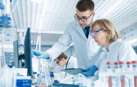 In Modern Laboratory Senior Female Scientist Has Discussion with Young Male Laboratory Assistant. He Shows Her Data Charts on a Clipboard, She Analyzes it and Enters It into Her Computer.