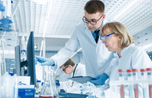 In Modern Laboratory Senior Female Scientist Has Discussion with Young Male Laboratory Assistant. He Shows Her Data Charts on a Clipboard, She Analyzes it and Enters It into Her Computer.