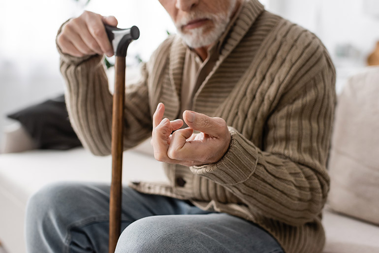 cropped view of senior man with parkinson disease holding walking cane while sitting on couch at home,stock image