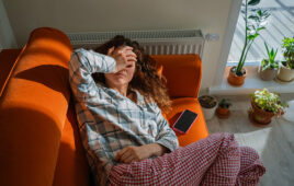 Woman feeling unwell and exhausted while lying on an orange sofa, covering her eyes with a hand, suffering from toxicosis, nausea, or a headache, indicating discomfort and illness