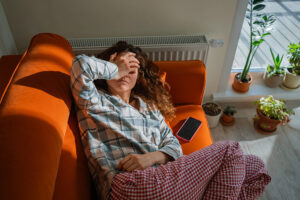 Woman feeling unwell and exhausted while lying on an orange sofa, covering her eyes with a hand, suffering from toxicosis, nausea, or a headache, indicating discomfort and illness