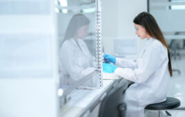 A pharmaceutical technician performs aseptic compounding inside a sterile biosafety cabinet, preparing a cell culture for biologic drug development under strict quality control conditions.