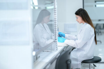 A pharmaceutical technician performs aseptic compounding inside a sterile biosafety cabinet, preparing a cell culture for biologic drug development under strict quality control conditions.