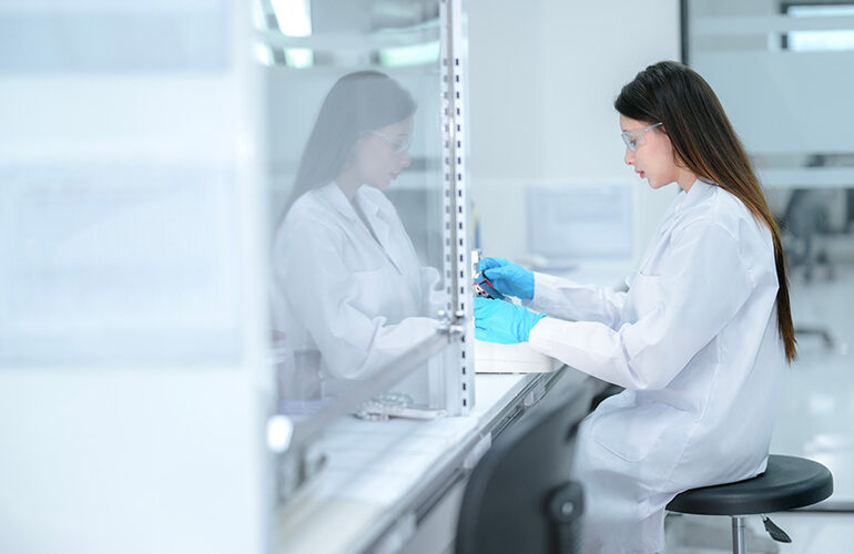 A pharmaceutical technician performs aseptic compounding inside a sterile biosafety cabinet, preparing a cell culture for biologic drug development under strict quality control conditions.