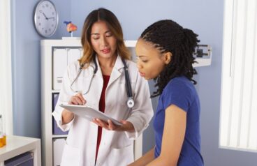 Physician in a white coat reviewing information on a tablet with a patient in an exam room.